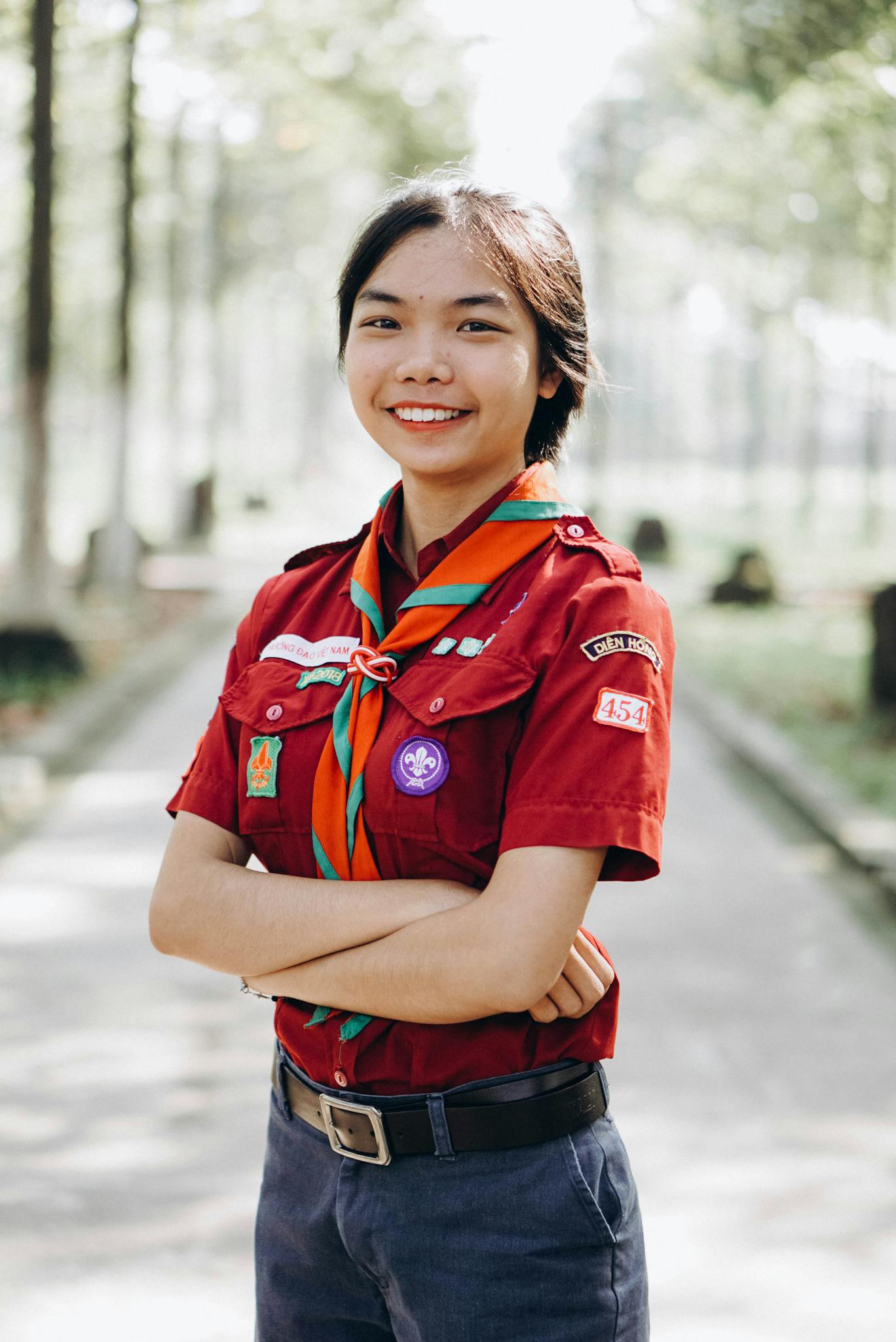 Girl in scout uniform smiling confidently outdoors with arms crossed.